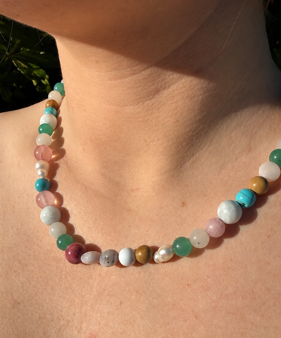 A close-up of a person's neck and upper chest wearing a beaded necklace made of round, polished stones in pastel pink, mint green, turquoise blue, white, and gray.